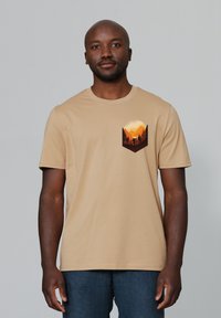 Beige katoenen T-shirt met een ronde halslijn, voorzien van een grafiek van bergen en bomen in warme kleuren op de borstzak. Korte mouwen.