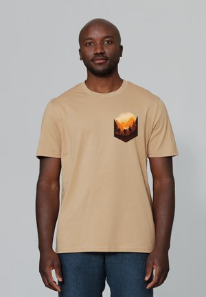 Tan cotton t-shirt with a round neckline, featuring a graphic of mountains and trees in warm colors on the chest pocket. Short sleeves.