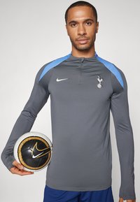 Graues langärmliges Sportshirt mit blauen Akzenten, mit einem Reißverschlusskragen und Vereinslogo, gehalten neben einem schwarz-weißen Nike-Fußball.