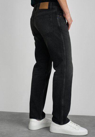 Des jeans en denim noir présentent une coupe droite, une texture délavée et une étiquette en cuir beige sur la taille. Ils sont associés à des baskets blanches.
