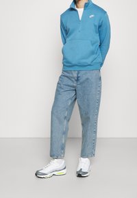 Sudadera de forro polar azul con cremallera y bolsillo frontal, combinada con jeans de pierna recta azul claro y zapatillas grises con acentos en neón.