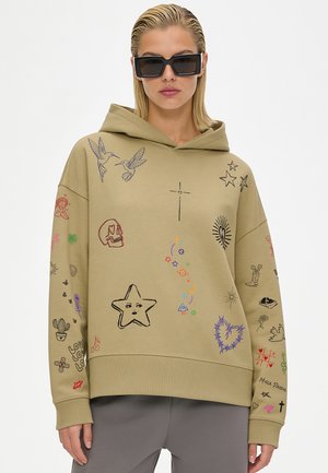 Woman wearing beige hoodie with colorful doodles including stars, birds, skull, heart, cross, and small symbols, paired with oversized black sunglasses.