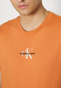 Oranžové bavlněné tričko s kulatým výstřihem a krátkými rukávy. Na hrudi výrazné bílé a černé logo "Calvin Klein Jeans".