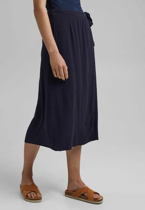Femme portant une jupe midi bleu marine foncé avec une ceinture à nouer, des sandales marron sans lacets et un haut sans manches bleu marine, debout sur un sol gris clair.