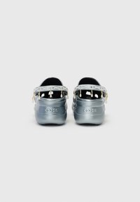 Des Crocs argentés avec des sangles à l'arrière, ornés d'une bande décorative, de petites perles pendantes et d'une surface texturée. Nom de la marque embossé sur le talon.