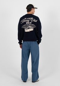 Homme portant une casquette noire et blanche, un pull marine avec le texte « In a Rush Racing Club Berlin » et un motif de voiture, ainsi qu'un pantalon bleu ample, debout de profil.