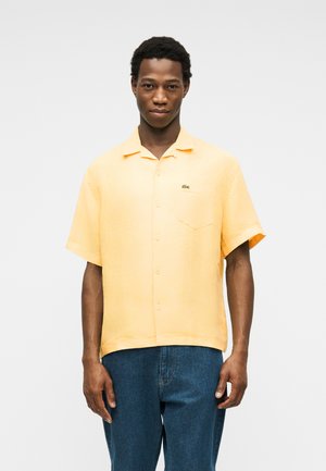 Jeune homme portant une chemise à manches courtes orange clair avec un petit logo sur la poche poitrine et un jean bleu.