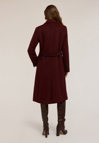 Cappotto in lana borgogna con vestibilità su misura, grande colletto e due bottoni posteriori. Presenta una cintura in vita e un design al di sotto del ginocchio.
