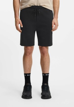 Pantaloni shorts neri con vita elastica e cordoncino; texture liscia; design semplice; indossati con sneakers nere e calzini con logo.