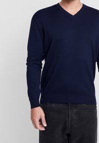 Marinblå V-ringad tröja i stickad tyg, med långa ärmar och ribbade muddar och nederkant. Bärs över svarta byxor, minimalistisk design.