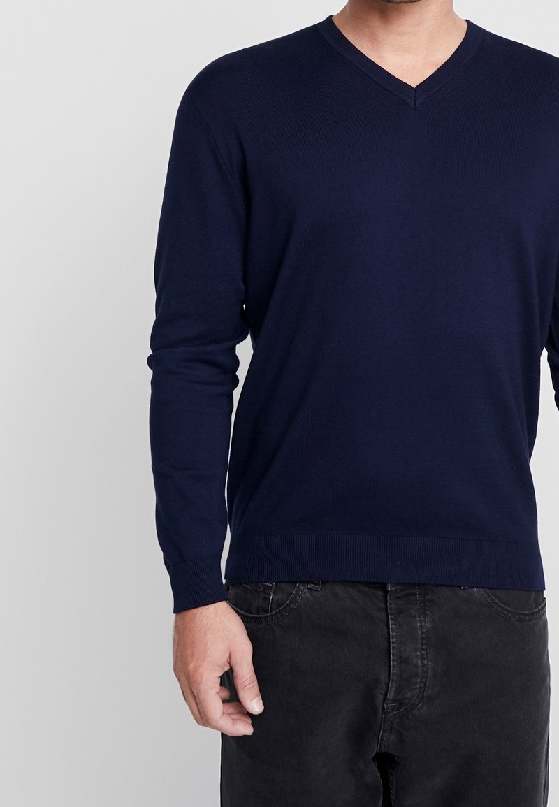 Marinblå V-ringad tröja i stickad tyg, med långa ärmar och ribbade muddar och nederkant. Bärs över svarta byxor, minimalistisk design.