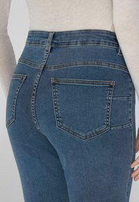 Tmavě modré džíny s vysokým pasem, zadním sedlem a dvěma zadními kapsami, které mají kontrastní prošití a kovové nýty.