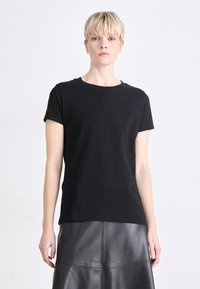 Melnā, īsām piedurknēm t-krekls ar apaļu kakla izgriezumu, ar smalkiem rotājumiem uz pleciem. Izgatavots no mīksta kokvilnas auduma.