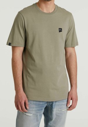 Mann trägt olivgrünes T-Shirt mit kurzen Ärmeln und kleinem schwarzem Logo auf der Brust und am linken Ärmel, kombiniert mit hellblauen zerrissenen Jeans.