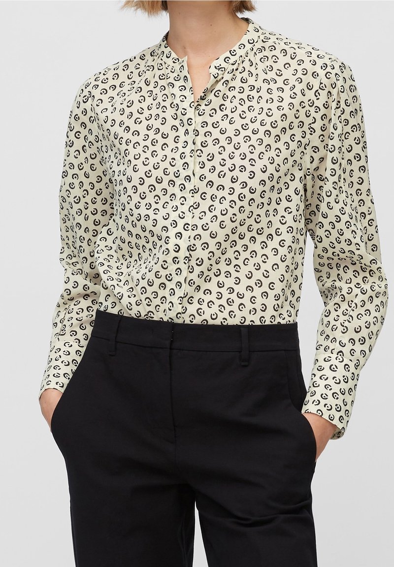 Personne portant un chemisier crème à motif circulaire noir et un pantalon noir, debout les mains dans les poches devant un fond uni.