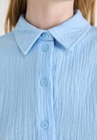 Camicia azzurro chiaro con colletto, realizzata in tessuto testurizzato, con cinque bottoni coordinati sul davanti e dettagli cuciti sul colletto.