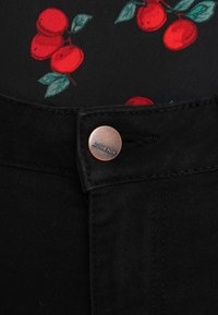 Pantalones de mezclilla negros con un botón de cobre grabado con "AUTÉNTICO". El fondo muestra una tela negra con cerezas rojas y hojas verdes.