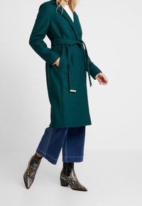 Manteau en laine teal avec un design enveloppant, taille ceinturée, poches latérales et manches longues. Associé à un jean évasé et des bottes en imitation serpent texturé.