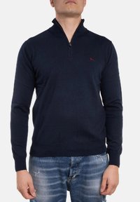 Pullover navy a maglia con colletto zip, dotato di polsini aderenti e un piccolo logo rosso sul petto, abbinato a jeans chiari strappati.