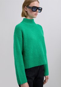 Grøn strikket sweater med høj krave, ribbet tekstur og kort pasform. Model bærer store sorte solbriller og mørke bukser.