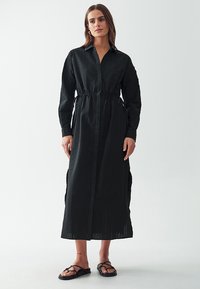 Abito nero a maniche lunghe in tessuto texture, colletto, vita con cordoncino e spacchi laterali, abbinato a semplici sandali neri.