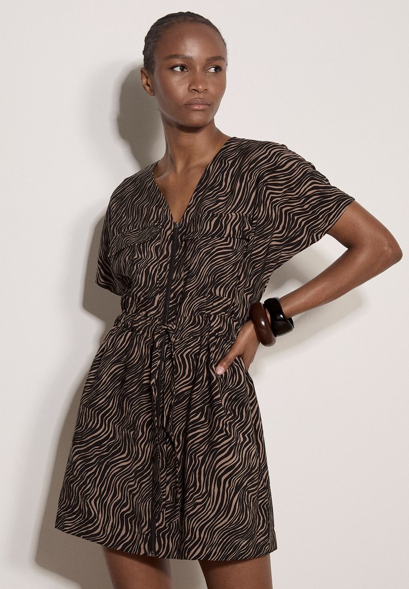 Femme portant une robe à imprimé zèbre marron et noir avec une fermeture éclair à l’avant et des bracelets noirs et marron, debout, une main sur la hanche.