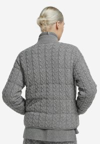 Pull gris en maille torsadée avec un col montant et des poignets côtelés. Le tissu présente un motif texturé, et la coupe est légèrement ajustée à la taille.