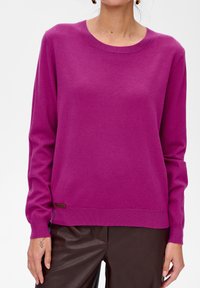 Fioletowy sweter dzianinowy z okrągłym dekoltem, długimi rękawami, ribbowanymi mankietami i dołem, z małą metką logotypu na dole. Połączony z brązowymi spodniami.