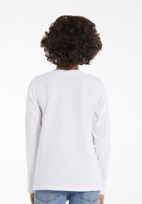 Langärmliges weißes Baumwollhemd von hinten gesehen, mit Rundhalsausschnitt und lockerer Passform, kombiniert mit hellblauen Jeans.