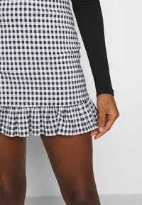 Schwarz-weißes Gingham-Minirock mit tailliertem Design und rüschsaum. Der Stoff ist leicht und hat eine glatte Textur. Hand, die den Rock hält.