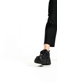 Persona staigā, valkājot melnus masīvus sporta apavus un melnas svītrains bikses, uz baltas grīdas pret vienkāršu baltu fonu.