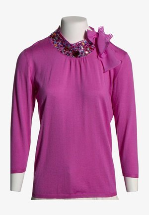 Haut à manches longues fuchsia avec encolure ornée de bijoux et grand nœud en tissu sur l'épaule gauche, présenté sur un mannequin blanc.
