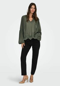 Donna che indossa una blusa trasparente verde oliva con orlo ricamato, jeans neri e scarpe con tacco beige, in piedi davanti a uno sfondo bianco.