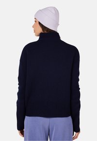 Navy blue knitted sweater with a high collar and ribbed hem. Model wears a light purple knit beanie, paired with light purple pants.