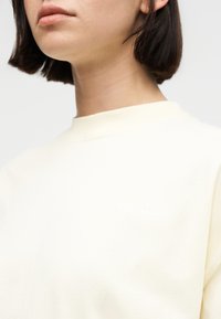 Gros plan d'une personne aux cheveux courts et foncés portant un haut à col rond de couleur crème sur un fond uni clair.