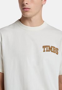 Tričko s krátkým rukávem v krémové barvě vyrobené z bavlny. Má kulatý výstřih a na levé hrudi je logo "TIMBS" v tučném oranžovém písmu.
