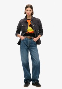 Chaqueta gris oscuro abotonada en el frente sobre una camiseta negra con un gráfico de atardecer. Unos vaqueros azules sueltos y zapatos negros completan el conjunto.