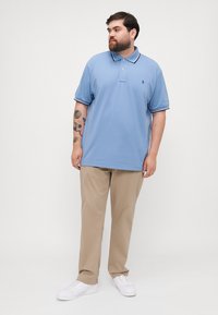 Ανοιχτό μπλε πουκάμισο πόλο με παλτό δύο κουμπιών και αντίθετο διακοσμητικό στα κολάρα, συνδυασμένο με μπεζ παντελόνια και λευκά αθλητικά παπούτσια.