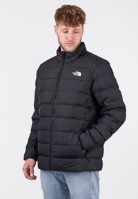 Giubbotto imbottito nero con zip frontale, dotato di un colletto alto e design a righe orizzontali. Logo sul petto a sinistra, texture del tessuto liscia.