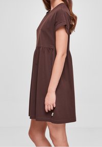 Hnědé bavlněné šaty s krátkými ohrnutými rukávy, kulatým výstřihem a nařaseným pasem, které se rozšiřují do uvolněného střihu do délky po kolena.