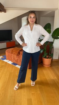 Blusa branca envolvente com mangas longas, combinada com calças largas azul-escuro e sandálias brancas de salto alto, contra um fundo de chão de madeira.