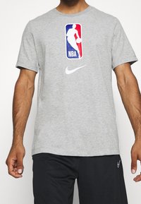 Camiseta de algodón gris con un gran logo de la NBA en azul y rojo, y un swoosh blanco de Nike en el pecho. Mangas cortas y cuello redondo.