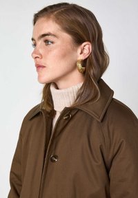 Giovane donna con capelli castano chiaro che indossa un cappotto marrone, un dolcevita beige e grandi orecchini a cerchio in oro testurizzato, rivolta verso sinistra.