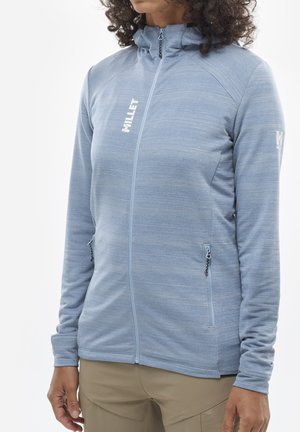 Woman wearing light blue full-zip jacket with high collar, zipped pockets, and "MILLET" logo on chest and sleeve.