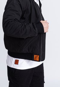 Homme portant une veste bomber noire avec une étiquette orange sur un t-shirt blanc et un pantalon noir, ajustant la fermeture éclair de la veste sur un fond uni.