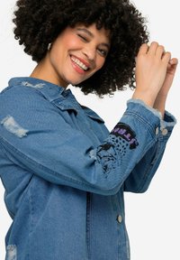 Jeansjacke in Hellblau mit abgenutzten Details. Verfügt über auffällige schwarze Grafik auf dem Ärmel, Druckknöpfe und ausgefranste Bündchen.