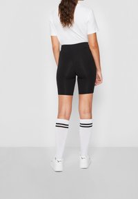 Kvinna som står och vänder ryggen till i svarta högmidjade cykelbyxor, vit kortärmad topp, vita knästrumpor med svarta ränder och vita sneakers.