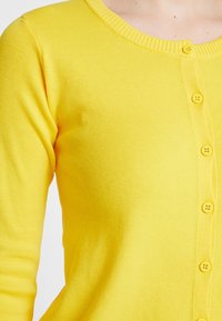 Yellow cardigan made from ribbed knit fabric. Features a round neckline and six matching yellow buttons down the front. Long sleeves.
