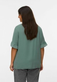 Blouse à manches courtes en vert délavé avec des ourlets de manches volants. Tissu léger avec une coupe ample, associé à un pantalon gris texturé.