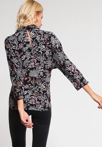 Schwarze Bluse mit floralen Mustern, dreiviertel langen Ärmeln, einem subtilen Schlüsselauschnitt am Rücken und einem gemischten Muster aus rosa und weißen botanischen Designs.